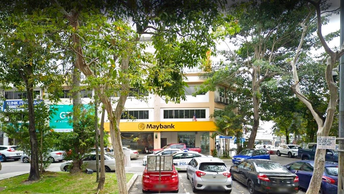 Pemandangan luar cawangan Maybank berjenama kuning yang terletak di Jalan Kuchai Lama, sebahagiannya dibingkai oleh pokok-pokok hijau yang rimbun. Beberapa buah kereta diletakkan di hadapan bangunan. Latar belakang menunjukkan kedai bersebelahan, termasuk sebuah klinik