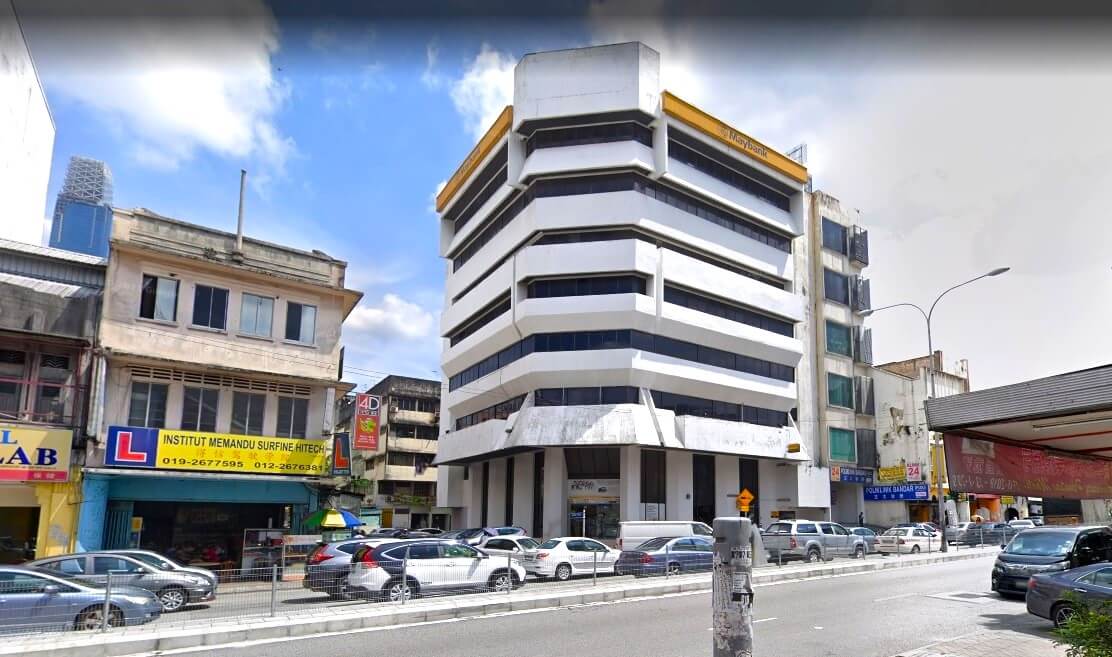 Bangunan cawangan Maybank Jalan Pudu, yang menampilkan fasad bersudut putih dan tingkap reben, terletak di jalan yang sibuk dengan kereta yang diletakkan