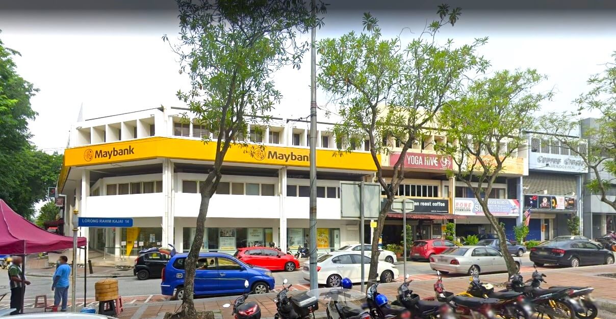 Pemandangan jalan bersudut lebar Maybank TTDI Branch di Taman Tun Dr. Ismail, memaparkan papan tanda kuning yang menonjol di seberang bangunan sudut tiga tingkat berwarna putih. Beberapa buah kereta dan motosikal diletakkan di sepanjang jalan di latar depan