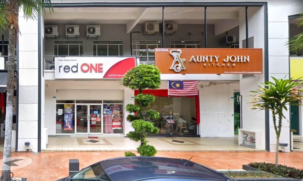 Pemandangan aras jalan bangunan komersial yang menampilkan redONE Network customer service center di sebelah kiri dan restoran Aunty John Kitchen di sebelah kanan