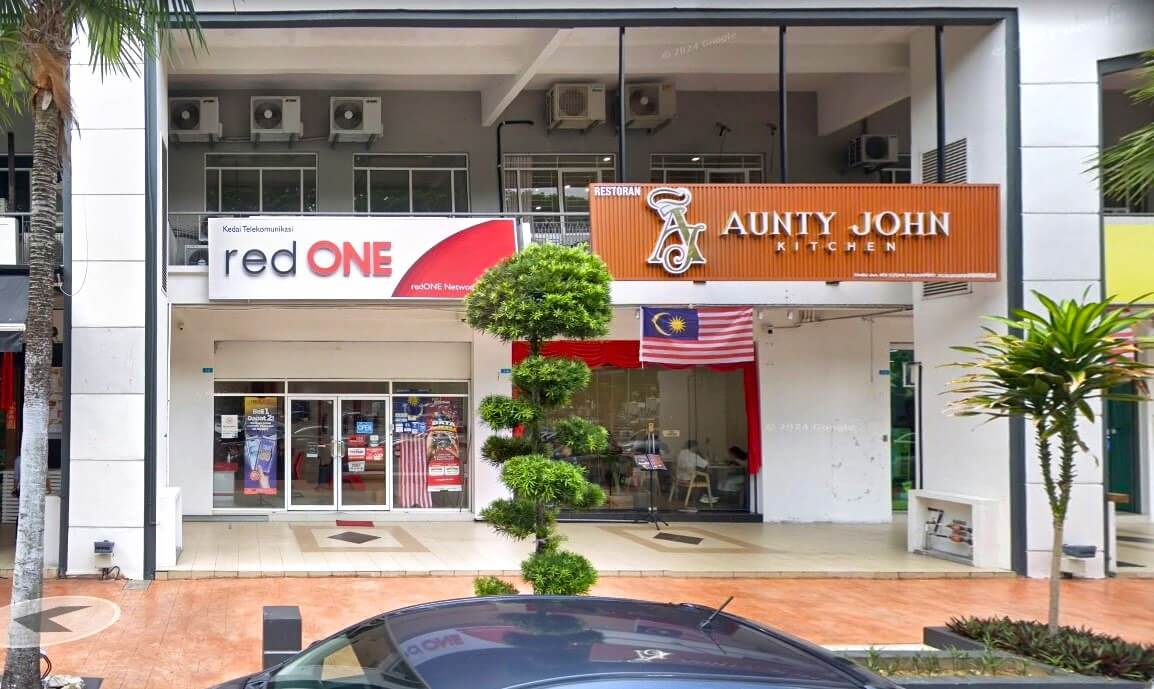 Pemandangan aras jalan bangunan komersial yang menampilkan redONE Network customer service center di sebelah kiri dan restoran Aunty John Kitchen di sebelah kanan