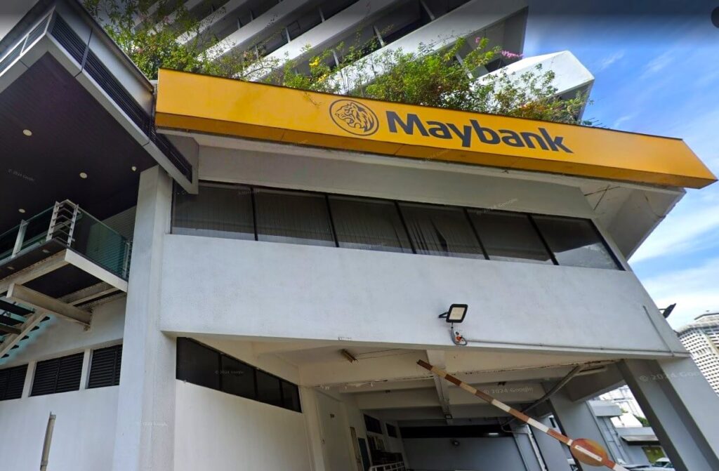 Pandangan luar cawangan Maybank yang terletak di bangunan putih bertingkat. Imej tersebut memaparkan papan tanda segi empat tepat kuning khas bank dengan logo harimau hitam dan teks "Maybank".