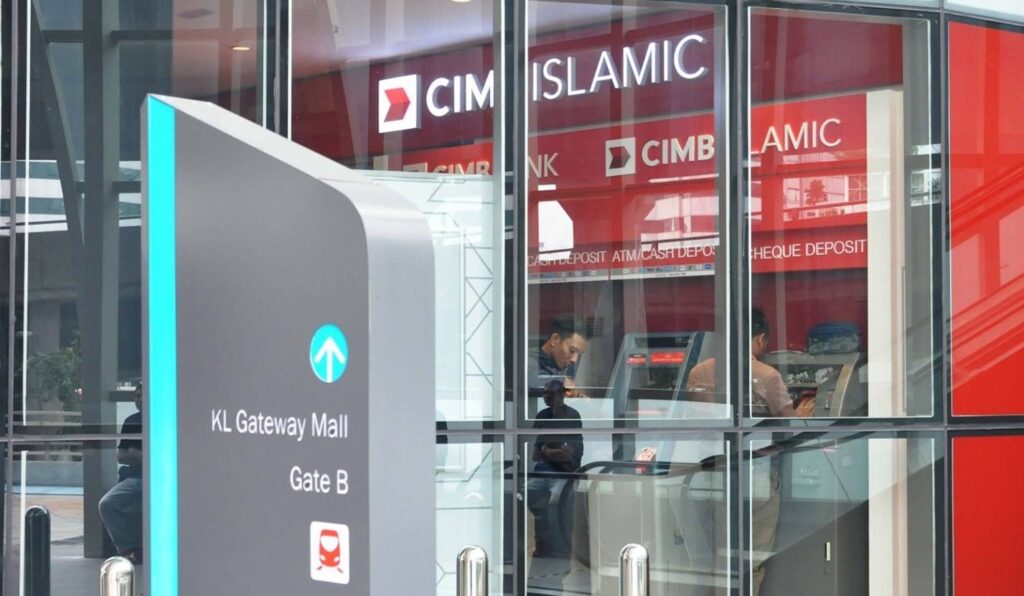 Gambar luaran cawangan CIMB Bank KL Gateway Mall (CIMB Islamic) di KL Gateway Mall menunjukkan kotak pameran kaca dengan logo merah dan putih. Di latar depan, tiang arah kelabu menghala ke "Gate B" di sebelah ikon kereta api. Di dalam kiosk berdinding kaca, dua lelaki dilihat menggunakan ATM
