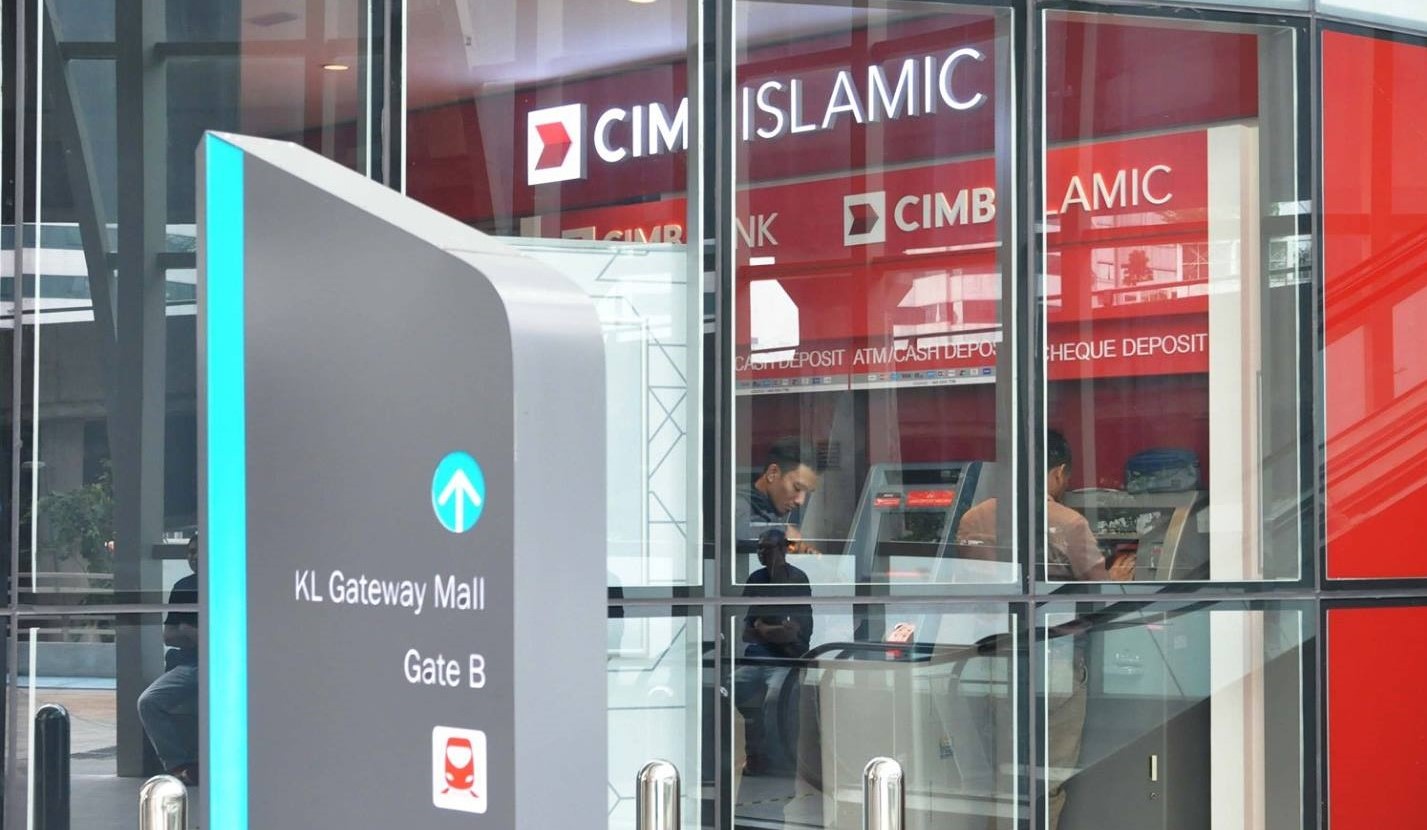 Gambar luaran cawangan CIMB Bank KL Gateway Mall (CIMB Islamic) di KL Gateway Mall menunjukkan kotak pameran kaca dengan logo merah dan putih. Di latar depan, tiang arah kelabu menghala ke "Gate B" di sebelah ikon kereta api. Di dalam kiosk berdinding kaca, dua lelaki dilihat menggunakan ATM