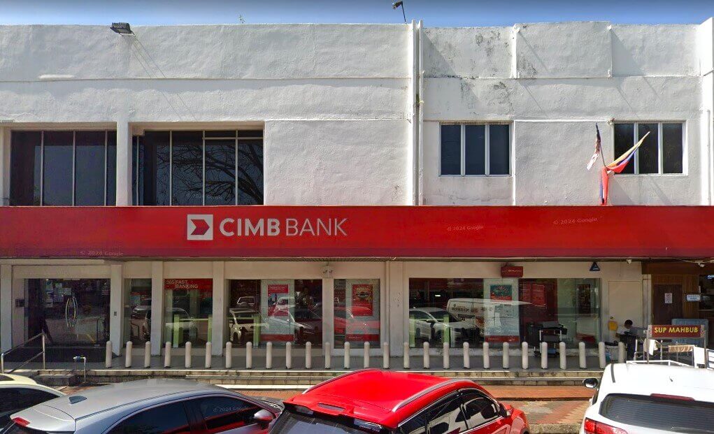 Pemandangan jalan cawangan CIMB Bank Lucky Garden Bangsar dua tingkat berwarna putih. Sepanduk merah panjang dengan logo "CIMB BANK" berwarna putih terbentang di hadapan kedai di atas tingkap kaca besar. Beberapa buah kereta, termasuk sebuah kereta berwarna merah yang menarik, diletakkan di latar depan, dan papan tanda "Sup Mahbub" kelihatan di sebelah kanan