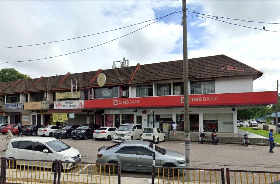 Pemandangan jalan bersudut lebar bangunan komersial dua tingkat berwarna putih yang menampilkan cawangan CIMB Bank dengan papan tanda merah yang menjadi ciri khasnya. Beberapa buah kereta diletakkan di hadapan bank dan kedai-kedai bersebelahan, termasuk sebuah restoran dan sebuah klinik