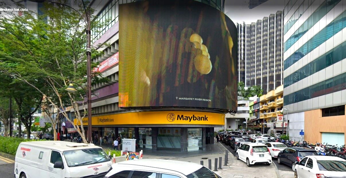 Pandangan aras jalan cawangan Maybank di persimpangan yang sibuk di Bukit Bintang, Kuala Lumpur, yang menampilkan papan iklan digital melengkung yang besar yang memaparkan iklan untuk Margaret River Region di atas kedai bank berwarna kuning terang