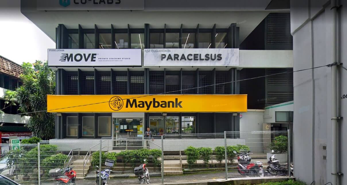 Bahagian luar cawangan Maybank yang terletak di sebuah bangunan bandar. Papan tanda berwarna kuning terang dengan logo dan nama Maybank merentangi bahagian tengah bingkai di atas pintu masuk. Di atas tebing, terdapat papan tanda untuk perniagaan lain, termasuk "MOVE Private Coaching Studio" dan "PARACELSUS."