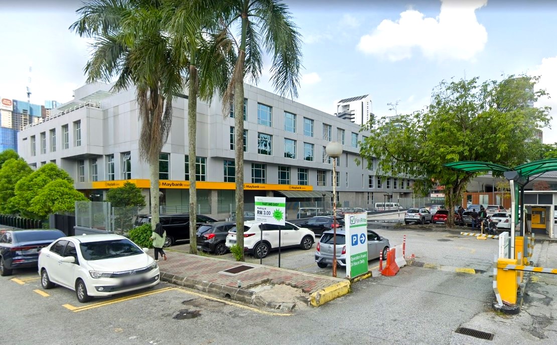 Pemandangan luar cawangan Maybank Islamic TTDI, menampilkan bangunan kelabu bertingkat moden dengan sepanduk korporat kuning. Di latar depan, terdapat tempat letak kereta ParkRite luar dengan beberapa kenderaan yang diletakkan, papan tanda kadar parkir hijau dan putih, dan pintu pagar penghalang masuk