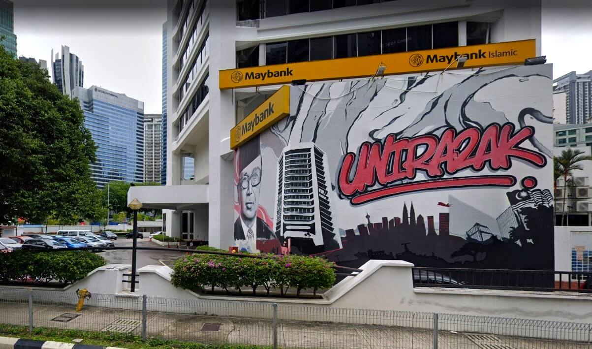 Sebuah mural di sisi cawangan Maybank Jalan Tun Razak di Kuala Lumpur, Malaysia. Karya seni bergaya grafiti yang besar itu menampilkan potret Tun Abdul Razak, latar langit Menara Berkembar Petronas, dan teks. Bangunan Maybank berwarna putih dengan papan tanda kuning, dan latar belakang bandar bertingkat tinggi moden