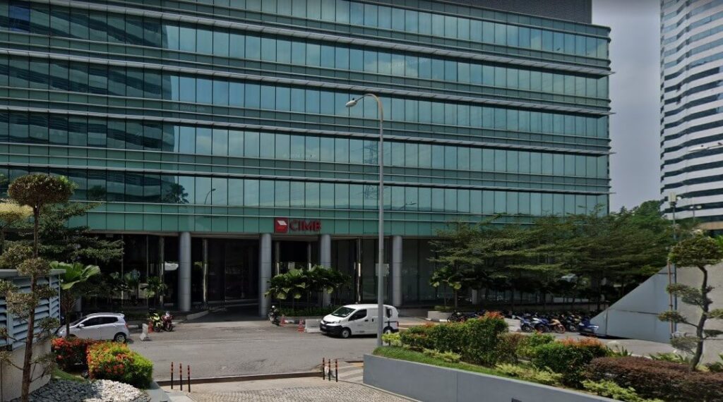Pemandangan aras jalan luar Wisma CIMB, sebuah bangunan pejabat moden bertingkat dengan fasad kaca hijau, menampilkan logo CIMB merah di atas pintu masuk dan kawasan parkir dengan kenderaan dan motosikal di latar depan
