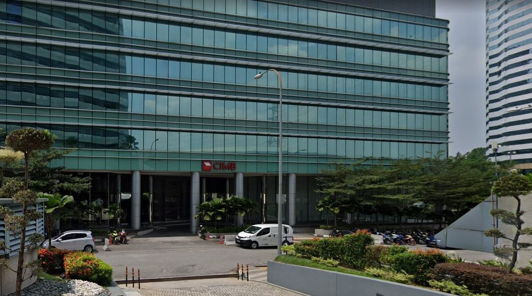 Pemandangan aras jalan luar Wisma CIMB, sebuah bangunan pejabat moden bertingkat dengan fasad kaca hijau, menampilkan logo CIMB merah di atas pintu masuk dan kawasan parkir dengan kenderaan dan motosikal di latar depan