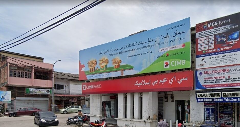 Gambar luar cawangan CIMB Bank yang terletak di barisan komersial. Sebuah papan iklan promosi biru dan hijau yang besar terletak di atas bangunan, memaparkan kempen "CIMB Bonus Booster" dengan teks dalam Melayu (tulisan Jawi) dan tulisan Latin