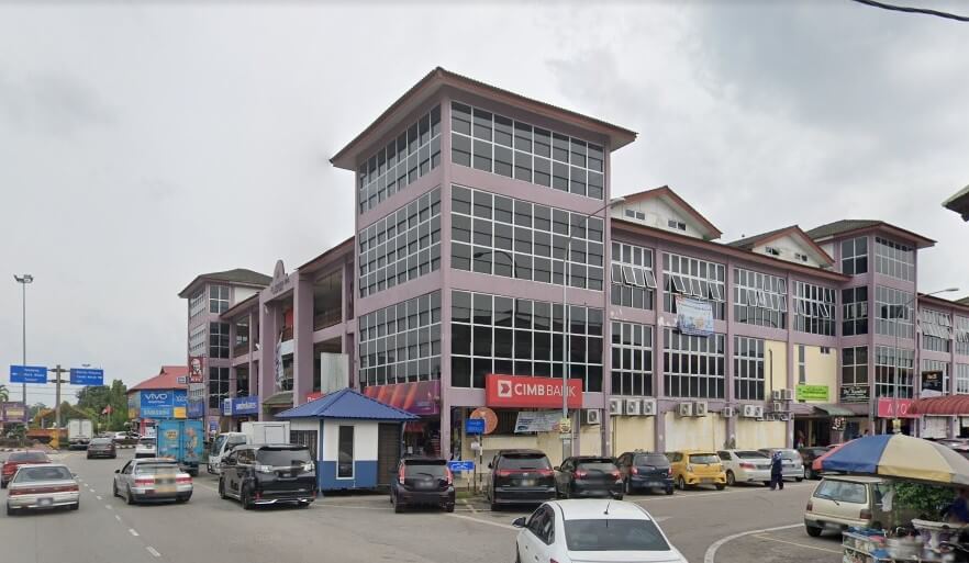 Sebuah bangunan komersial berwarna ungu bertingkat di sudut jalan yang memaparkan papan tanda CIMB Bank merah yang menonjol. Jalan di hadapan sibuk dengan pelbagai kereta yang diletakkan dan bergerak, sebuah pondok pengawal kecil berwarna biru dan putih, dan gerai makanan jalanan di latar depan