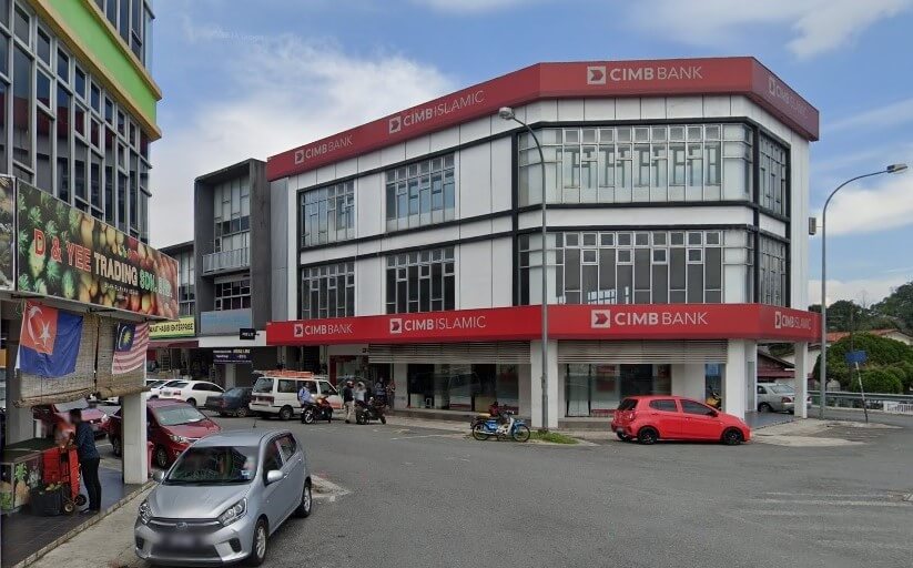 Sebuah bangunan sudut tiga tingkat cawangan CIMB Bank dan CIMB Islamic di Senai, Malaysia, yang menampilkan pelapisan putih dan papan tanda merah yang menonjol. Beberapa buah kereta dan motosikal diletakkan di sepanjang jalan di hadapan bank