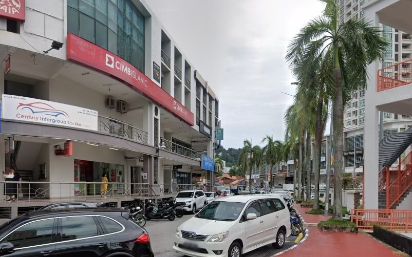 Pemandangan luar cawangan CIMB Bank Prima Tanjung yang terletak di bangunan komersial bertingkat di Prima Tanjung, Pulau Pinang. Pemandangan aras jalan menunjukkan beberapa kereta dan motosikal yang diletakkan di sepanjang jalan yang melengkung, dengan pokok palma berbaris di laluan pejalan kaki berturap merah