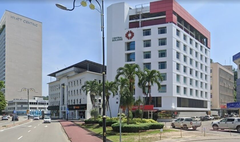 Jalan Central Building di Kota Kinabalu, menampilkan cawangan CIMB Bank di tingkat bawah. Bangunan bertingkat putih itu terletak di sudut bersebelahan jalan berturap dengan lalu lintas yang lancar dan pokok palma. Di latar belakang, hotel Hyatt Centric dan bangunan komersial lain kelihatan