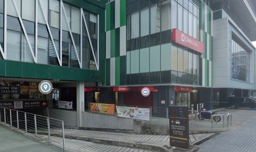 Pandangan luar cawangan CIMB Bank yang terletak di Riverson, menunjukkan bangunan bertingkat moden dengan panel fasad hijau dan putih. Papan tanda merah bank itu kelihatan di sudut bangunan, di atas pintu masuk aras tanah