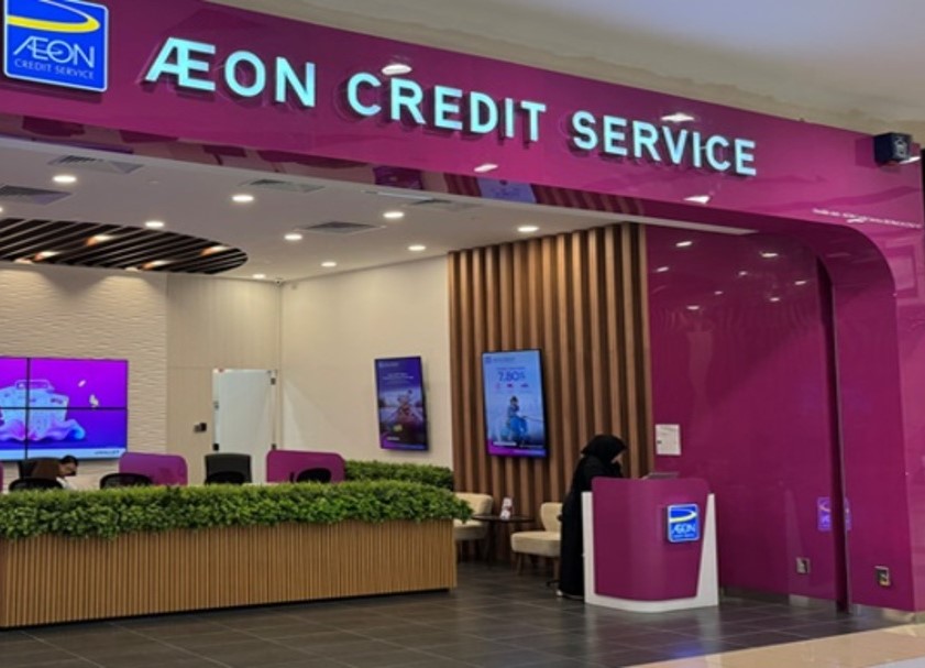 Foto interior yang menunjukkan kotak paparan dan bahagian dalam cawangan AEON Credit Service. Papan tanda yang menarik perhatian di atas pintu masuk ialah magenta terang dengan teks putih. Di dalam, terdapat kaunter perkhidmatan di sebelah kanan di mana seseorang berpakaian gelap dan memakai tudung berdiri, dan kaunter kayu panjang yang dihiasi dengan tumbuhan hijau tiruan di sebelah kiri