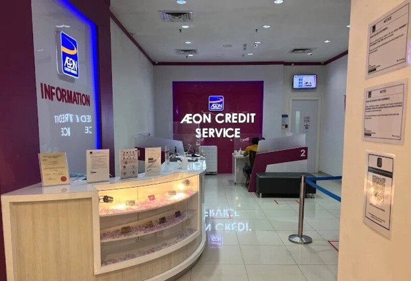 Pandangan dalaman cawangan AEON Credit Service dengan kaunter kaca melengkung di latar depan, dengan tanda "INFORMATION" di dinding di belakangnya, di sebelah kiri. Panel dinding besar di tengah latar belakang memaparkan "AEON CREDIT SERVICE" dalam teks putih pada latar belakang merah jambu/ungu gelap