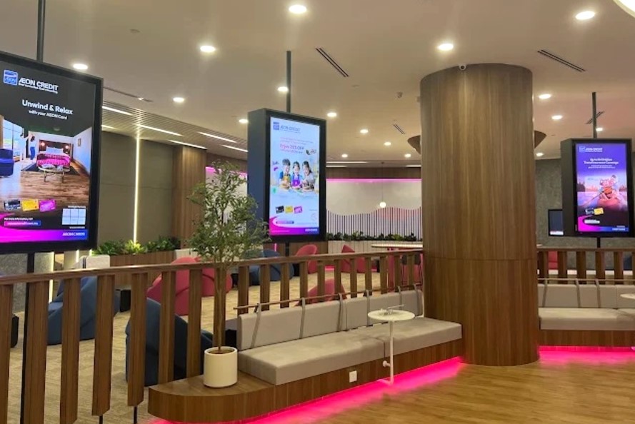 Bahagian dalam AEON Credit Bukit Mertajam Flagship Branch, menampilkan ruang menunggu moden dengan bangku kayu kelabu muda, pagar kayu rendah, skrin pengiklanan digital tinggi yang memaparkan promosi, dan ruang istirahat dengan pencahayaan aksen merah jambu di sepanjang lantai