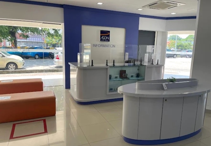 Bahagian dalam lobi pejabat AEON Credit Service, menampilkan meja maklumat berwarna putih dengan skrin plexiglass pelindung pada dinding aksen biru dengan logo syarikat. Di sebelah kiri terdapat bangku menunggu berwarna oren, dan di latar depan terdapat meja paparan bujur putih. Tingkap-tingkap besar menghadap ke kawasan tempat letak kereta
