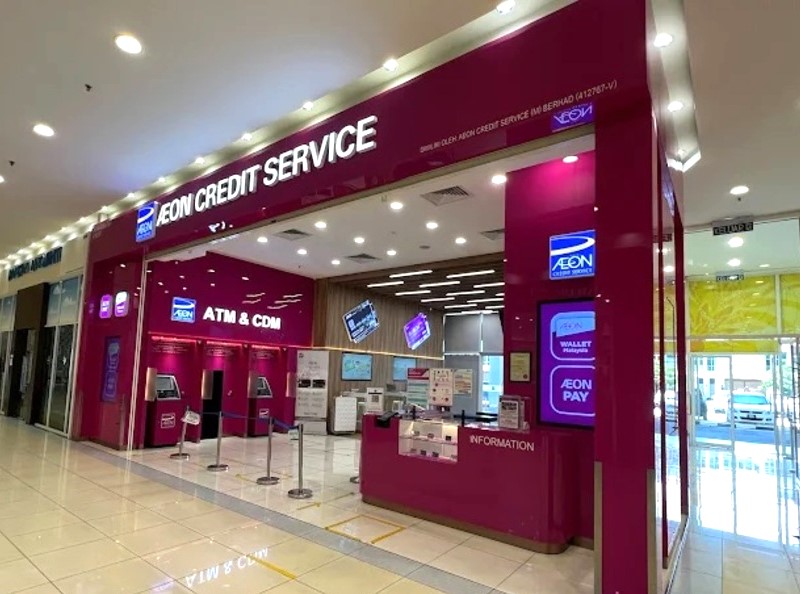 Etalase berwarna merah jambu dan putih terang untuk "AEON CREDIT SERVICE" yang terletak di dalam pusat beli-belah, menampilkan kaunter "INFORMATION", beberapa skrin yang memaparkan perkhidmatan seperti "AEON PAY" dan "Wallet Malaysia," serta bahagian untuk "ATM & CDM."