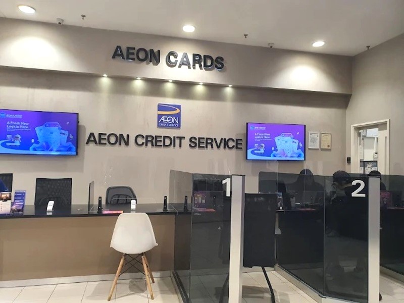 Pandangan dalaman kaunter AEON Credit Service di AEON Credit Queensbay berlabel 'AEON CARDS' dan 'AEON CREDIT SERVICE' pada dinding berwarna krim, menampilkan dua skrin digital dan meja khidmat pelanggan dengan partition lutsinar