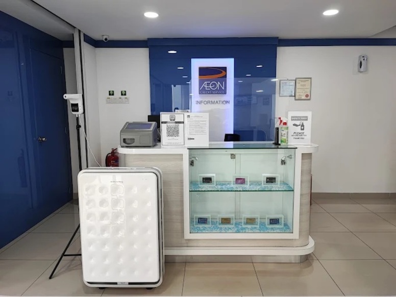 Bahagian dalam cawangan AEON Credit Services menampilkan meja maklumat berwarna putih dengan kotak paparan kaca yang mengandungi kad kredit. Pembersih udara putih besar dan pengimbas suhu berdiri di sebelah kiri, manakala kaunter dilengkapi dengan perisai akrilik, pembersih tangan dan papan kod QR