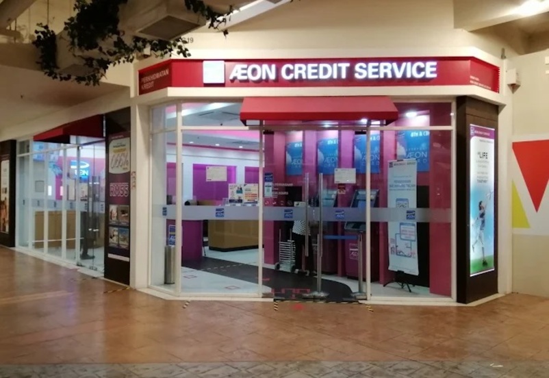 Pemandangan luar pejabat "AEON Credit Service" dengan pencahayaan dalaman merah jambu dan putih, terletak di dalam pusat membeli-belah atau bangunan. Pintu masuk ialah pintu kaca di bawah kanopi merah, dan tandanya ialah teks putih pada latar belakang merah
