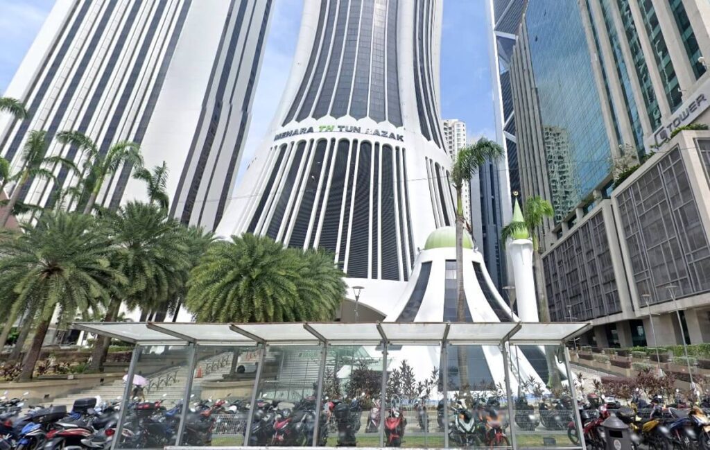 Pemandangan sudut rendah bangunan pencakar langit berbelang putih dan hitam, Menara TH Tun Razak, menjulang tinggi di atas kawasan parkir motosikal beratap kaca dengan pokok palma di latar depan, dan bangunan moden bersebelahan lain di Kuala Lumpur