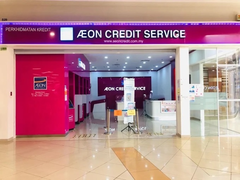 Etalase magenta yang terang untuk "AEON CREDIT SERVICE" kelihatan di dalam pusat membeli-belah, menampilkan papan tanda bercahaya terang di atas pintu masuk dan dinding kaca. Bahagian dalamannya, juga magenta, termasuk meja perkhidmatan dan meja maklumat