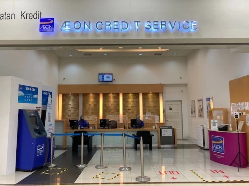 Interior pejabat AEON Credit Metro Prima, menunjukkan papan tanda terang bertulis "AEON CREDIT SERVICE" di atas meja yang disediakan oleh dua pekerja. Di sebelah kiri terdapat ATM/kiosk layan diri berwarna biru. Lantai mempunyai anak panah arah untuk "IN" dan "OUT"
