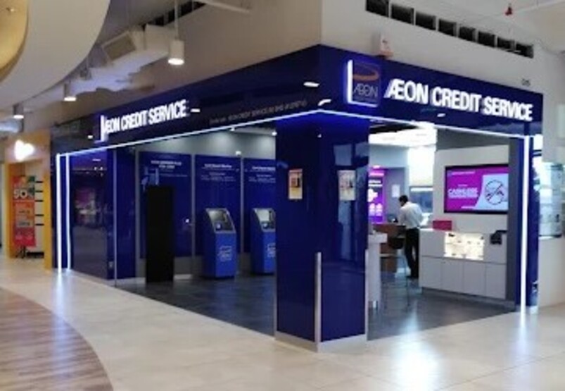 Gambar dalaman cawangan AEON Credit Taman Maluri. Bahagian hadapan kedai bercahaya terang dengan aksen biru dan putih, menampilkan logo "AEON CREDIT SERVICE" di bahagian hadapan. Di dalam pintu masuk, terdapat beberapa kiosk layan diri dan kaunter perkhidmatan dengan seorang kakitangan kelihatan di latar belakang
