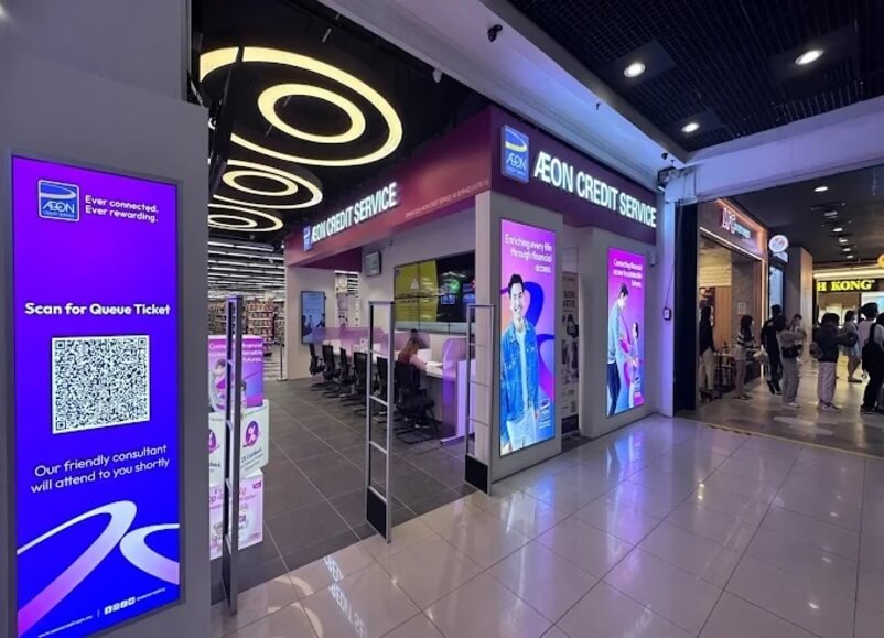 Cawangan AEON Credit Service yang cerah dan moden yang terletak di dalam pusat beli-belah, menampilkan penjenamaan ungu, skrin paparan digital dengan imej promosi orang, kod QR untuk tiket giliran, dan deretan meja perkhidmatan di dalamnya