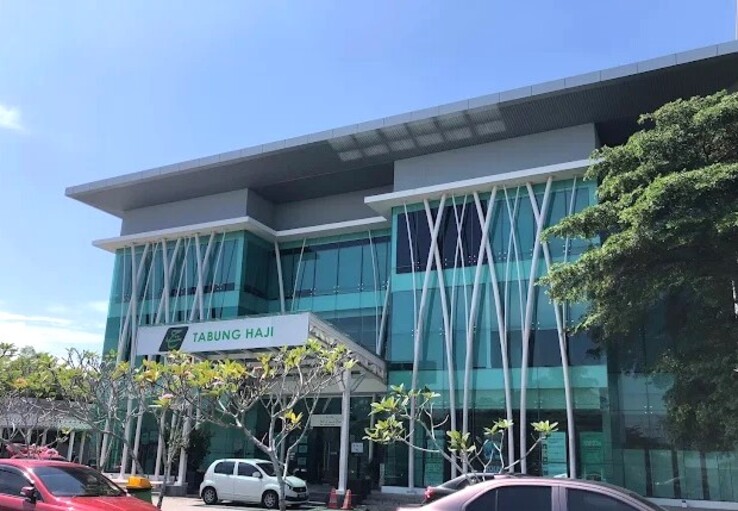 Sebuah bangunan kaca tiga tingkat moden dengan tiang putih dihiasi ukiran palang dan papan tanda yang tertulis "TABUNG HAJI" di bawah langit biru yang cerah