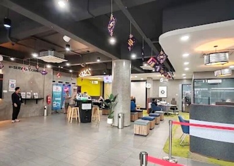 Bahagian dalaman moden pelan terbuka pejabat KWSP (Kumpulan Wang Simpanan Pekerja) di Kluang. Ruang ini menampilkan siling kelabu gaya perindustrian dengan saluran terdedah, tiang konkrit, dan "E-Service Corner." Kakitangan dan pelanggan boleh dilihat berhampiran kiosk perkhidmatan dan ruang tempat duduk, yang merangkumi bangku kayu dan kerusi berlapis kain biru