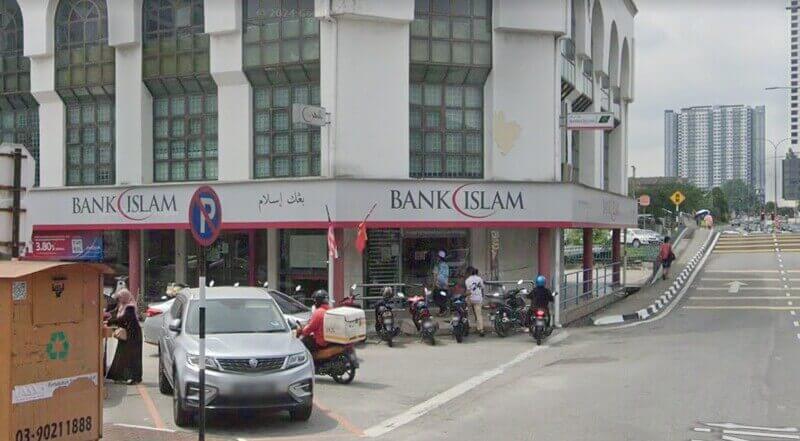 Pandangan luar cawangan Bank Islam Malaysia Berhad di sudut Kajang, Malaysia, menampilkan papan tanda putih dengan aksen merah, motosikal yang diletakkan di hadapan, dan pemandangan jalan dengan kenderaan yang lalu lalang