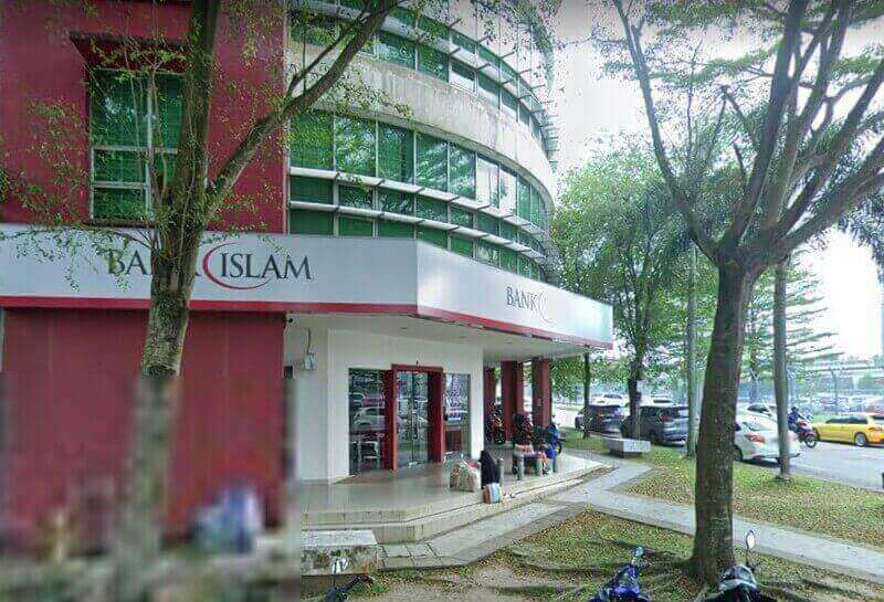 Pemandangan luar cawangan Bank Islam Malaysia Berhad di Putra Heights, menampilkan bangunan melengkung berwarna merah dan putih dengan tingkap kaca besar, pokok-pokok hijau di latar depan, dan jalan dengan kereta yang diletakkan di latar belakang