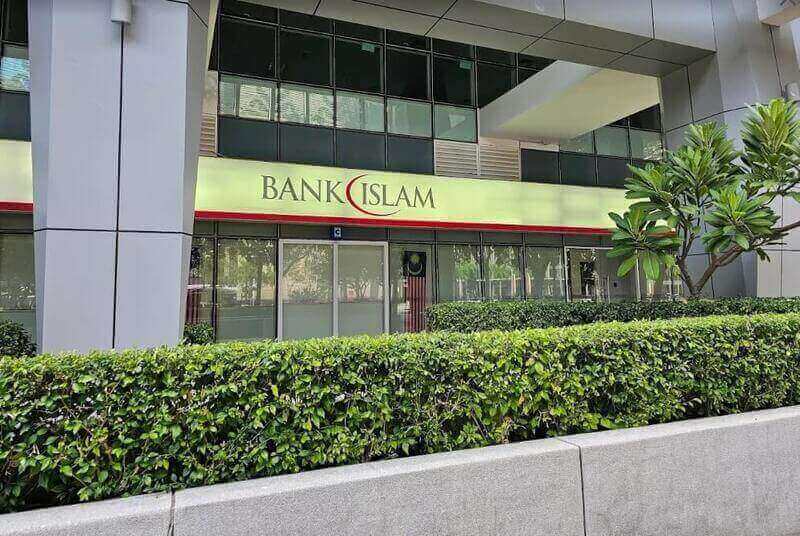 Pandangan luar cawangan Bank Islam Malaysia Berhad di Putrajaya, memaparkan papan tanda korporat hijau dan merah di atas pintu masuk kaca, sebahagiannya dikaburkan oleh pagar hijau yang dihias rapi