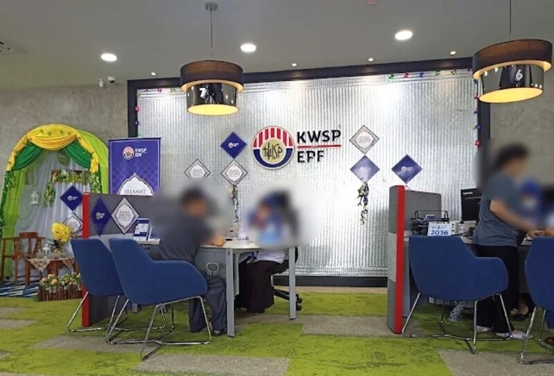 Pemandangan dalaman pejabat cawangan perkhidmatan KWSP (EPF) di Malaysia. Pejabat ini menampilkan bahagian dalaman moden dengan permaidani hijau, kerusi biru dan kaunter perkhidmatan di mana kakitangan sedang membantu pelanggan. Dinding bertekstur yang besar memaparkan logo "KWSP EPF" di samping hiasan Hari Raya yang meriah, termasuk gerbang berlapis hijau dan kuning