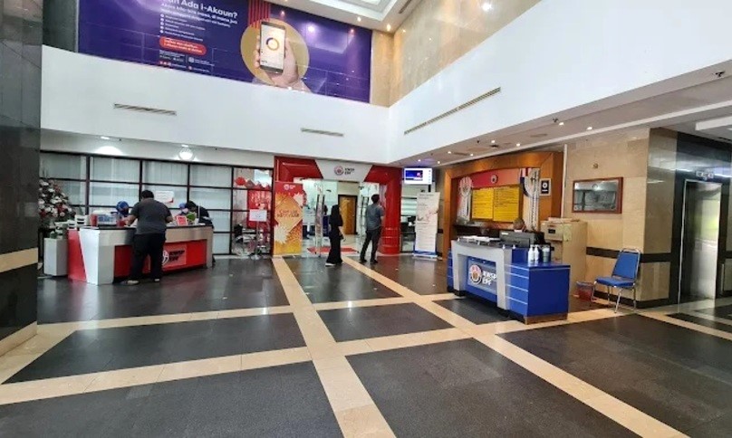 Pemandangan dalaman lobi pejabat KWSP (EPF) di Johor Bahru. Dewan yang luas dan terang benderang ini menampilkan lantai jubin berkilat dan siling tinggi. Di sebelah kiri terdapat kaunter perkhidmatan merah dengan kakitangan, dan di sebelah kanan terdapat meja maklumat biru. Di latar belakang, gerbang merah menuju ke kawasan pejabat yang lebih jauh, sementara sepanduk promosi ungu besar untuk aplikasi mudah alih "i-Akaun" dipaparkan di dinding di atas