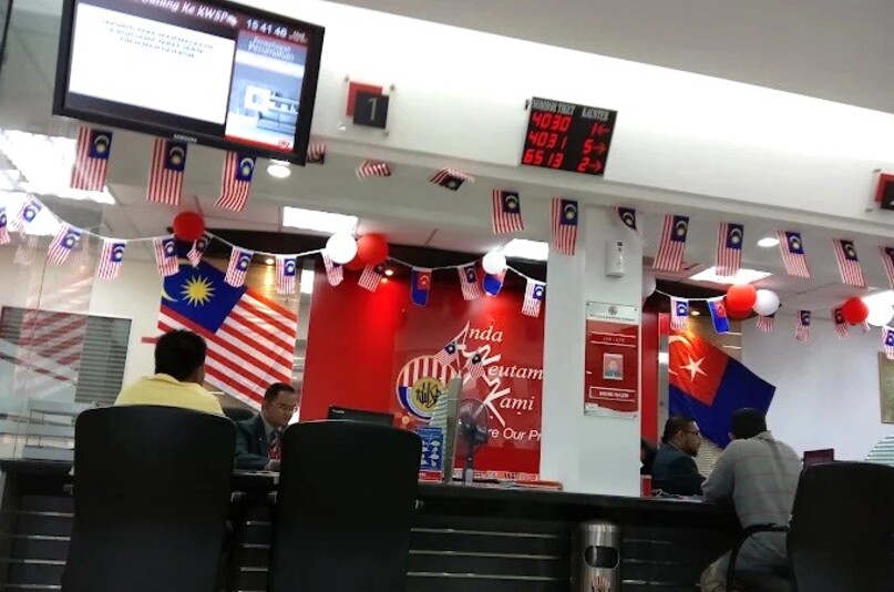 Bahagian dalam pejabat KWSP (Kumpulan Wang Simpanan Pekerja) di Malaysia, dihiasi dengan pelbagai bendera Malaysia (Jalur Gemilang) dan belon merah putih. Pelanggan duduk di kaunter perkhidmatan yang panjang dan dilayan oleh kakitangan