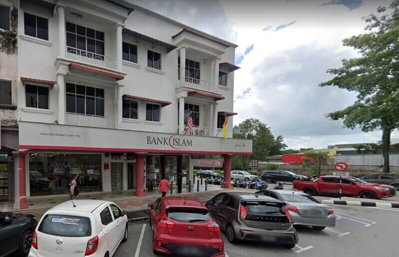 Pandangan luar cawangan Bank Islam Malaysia Berhad tiga tingkat di Bandar Baru Nilai, menampilkan bahagian hadapan putih dengan tiang merah, tempat letak kereta aras jalan dengan beberapa buah kereta, dan logo bank dipaparkan dengan jelas di atas pintu masuk