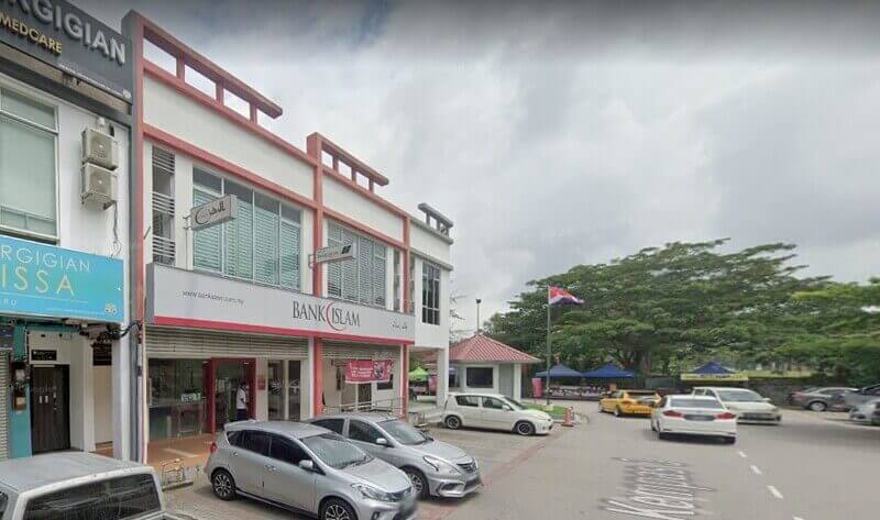 Pandangan luar cawangan Bank Islam Malaysia Berhad di Bandar Penawar, Malaysia, yang terletak di bangunan komersial dua tingkat berwarna putih dan merah dengan kereta diletakkan di hadapan dan bendera negeri Malaysia kelihatan di latar belakang