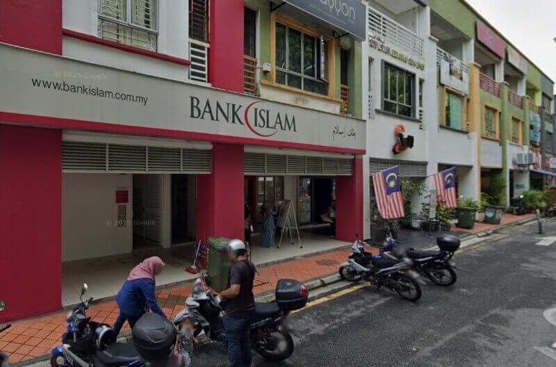 Pemandangan jalan cawangan Bank Islam di Malaysia, menampilkan kedai berwarna merah dan putih dengan logo dan laman web bank. Beberapa buah motosikal diletakkan di kaki lima di hadapan, dan bendera Malaysia dipamerkan di bangunan bersebelahan
