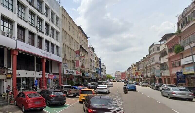 Pemandangan jalan raya komersial yang sibuk di Kluang, Malaysia, menampilkan cawangan Bank Islam Malaysia Berhad Kluang di sebelah kiri, lot kedai bertingkat dan kereta yang diletakkan di tepi jalan di bawah langit mendung