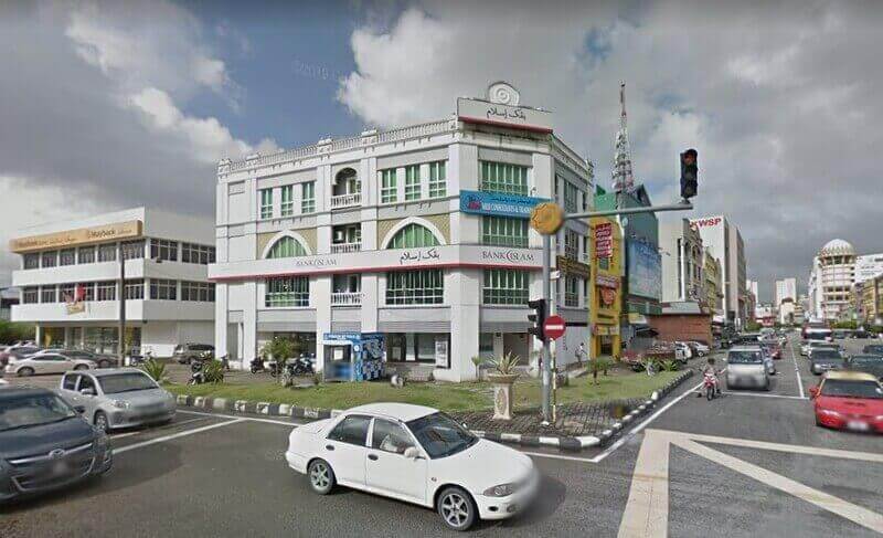 Sebuah bangunan Bank Islam Malaysia Berhad Padang Garong putih tiga tingkat dengan tingkap melengkung di sudut jalan di Padang Garong, Malaysia, memaparkan papan tanda Arab dan Inggeris di bawah langit mendung dengan lalu lintas bandar di latar depan