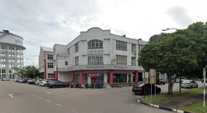 Sebuah bangunan sudut tiga tingkat yang menampilkan cawangan Bank Islam dengan dinding putih, tiang merah jambu, dan beberapa buah kereta serta motosikal yang diletakkan di hadapan di bawah langit mendung