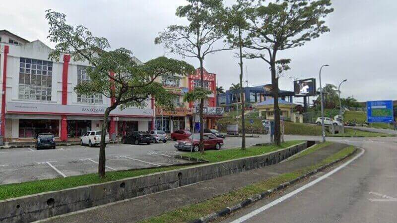 Pemandangan aras jalan cawangan Bank Islam Malaysia Berhad di Tampoi, menampilkan kedai putih dengan aksen merah, kawasan parkir berturap dan kehijauan di sekelilingnya di bawah langit mendung